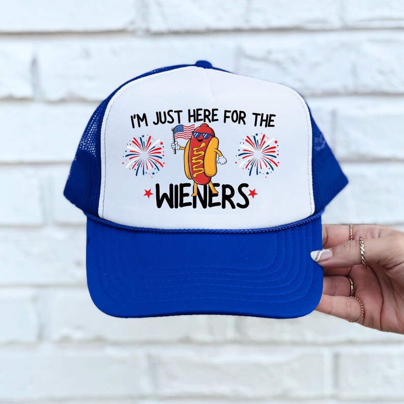 Puede incluir: Una gorra de camionero azul y blanca con una bandera estadounidense roja, blanca y azul y un perro caliente de dibujos animados que sostiene una bandera estadounidense m&aacute;s peque&ntilde;a. El texto en la gorra dice "I'm just here for the wieners".