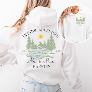 May include: White hoodie with a nature-themed design on the back, including mountains, trees, a river, and a sun. The text says "LIFETIME ADVENTURE" and "Mr. & Mrs. BARSTEN", with "EST. 2026". A smaller version of the design is on the front.