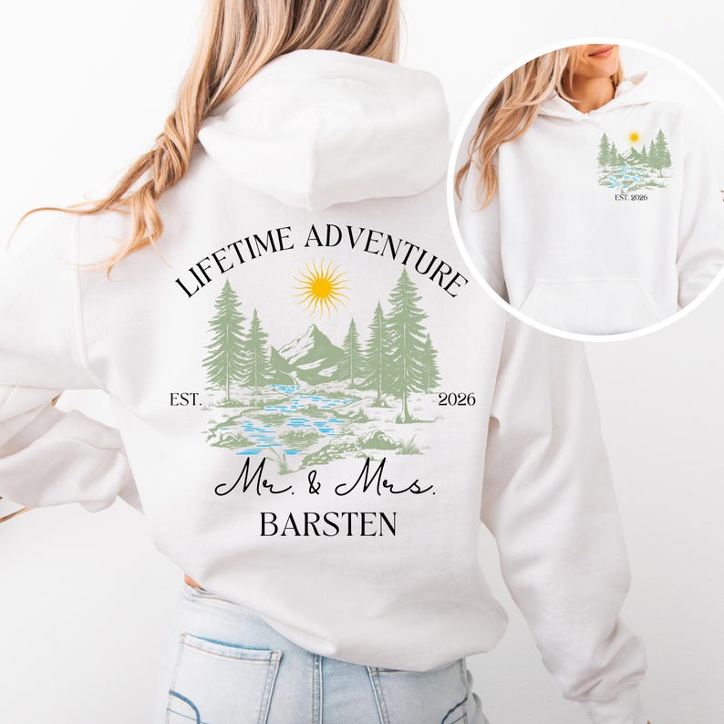 May include: White hoodie with a nature-themed design on the back, including mountains, trees, a river, and a sun. The text says "LIFETIME ADVENTURE" and "Mr. & Mrs. BARSTEN", with "EST. 2026". A smaller version of the design is on the front.