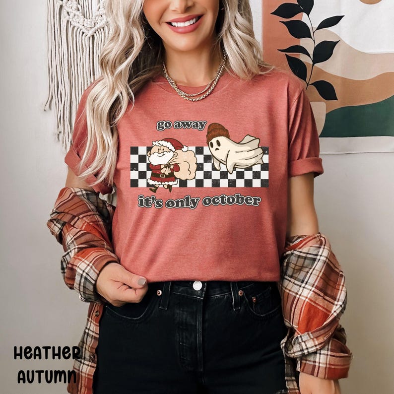 May include: A red t-shirt with a graphic of a ghost kicking Santa Claus away with the text "go away it's only october" on a checkered background.