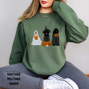 May include: Heather military green sweatshirt featuring three dogs dressed in Halloween costumes. One dog is in a ghost costume, another in a witch costume, and the third in a cloak. Each dog is holding a pumpkin-shaped treat bag.