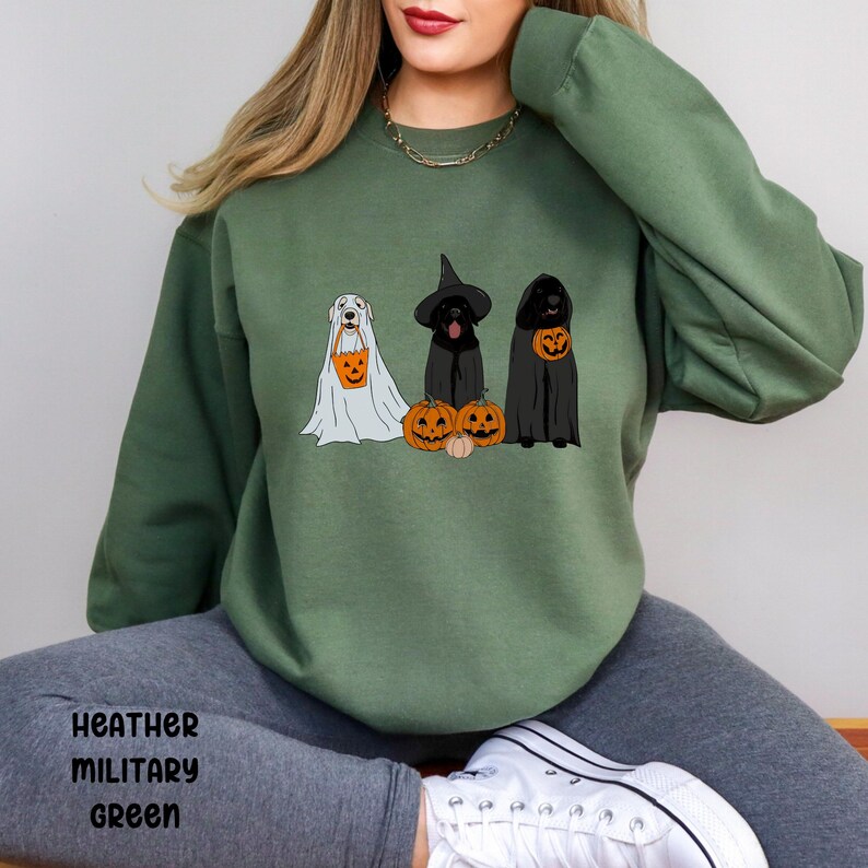 May include: Heather military green sweatshirt featuring three dogs dressed in Halloween costumes. One dog is in a ghost costume, another in a witch costume, and the third in a cloak. Each dog is holding a pumpkin-shaped treat bag.
