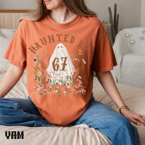 Puede incluir: Camiseta naranja con un dise&ntilde;o de fantasma, el n&uacute;mero 67 y el texto "HAUNTED BY", rodeado de flores. Una prenda informal, ideal para Halloween o eventos tem&aacute;ticos.