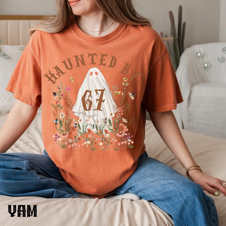 Puede incluir: Camiseta naranja con un dise&ntilde;o de fantasma, el n&uacute;mero 67 y el texto "HAUNTED BY", rodeado de flores. Una prenda informal, ideal para Halloween o eventos tem&aacute;ticos.
