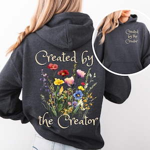 May include: Dark gray hoodie with a floral design and the text "Created by the Creator." The design features colorful flowers in red, pink, yellow, and blue. The same text is on the hood. A casual and comfortable apparel item.