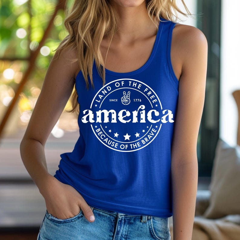 Pode incluir: Uma regata azul royal com a palavra "america" em branco, rodeada por um design circular que diz "Land of the Free Since 1776 Because of the Brave". O design inclui um sinal de paz.