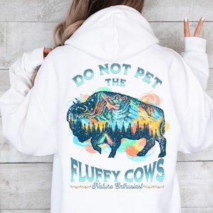 May include: White hoodie with a blue and green illustration of a bison with mountains and trees inside. The text "Do Not Pet The Fluffy Cows" is printed on the hoodie with the words "Nature Enthusiast" underneath.