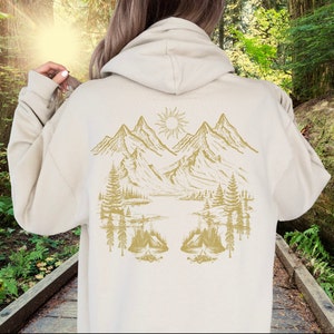 May include: A cream-coloured hoodie with a gold mountain scene on the back. The scene includes mountains, trees, a lake, and a sun.