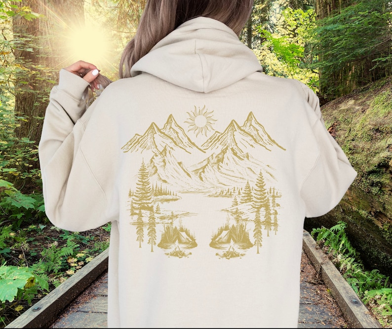 May include: A cream-coloured hoodie with a gold mountain scene on the back. The scene includes mountains, trees, a lake, and a sun.