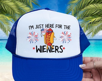 May include: A white and blue trucker hat with a red, white, and blue hot dog wearing sunglasses and holding a small American flag. The hat has the text "I'm just here for the WIENERS" with red stars on either side of the word "WIENERS".