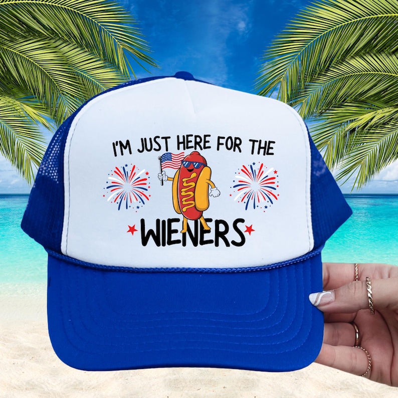Puede incluir: Una gorra de camionero blanca y azul con un perrito caliente rojo, blanco y azul que lleva gafas de sol y sostiene una peque&ntilde;a bandera estadounidense. La gorra tiene el texto "I'm just here for the WIENERS" con estrellas rojas a cada lado de la palabra "WIENERS".