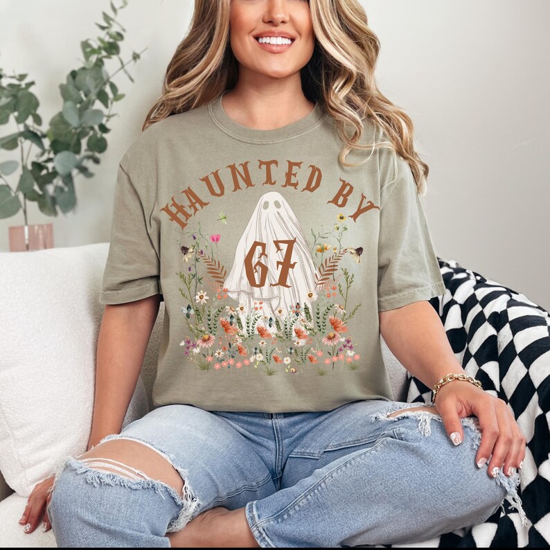 Puede incluir: Camiseta verde oliva con el texto "HAUNTED BY 67" en letras marrones. El dise&ntilde;o presenta una figura de fantasma blanco con el n&uacute;mero 67, rodeada de flores y follaje coloridos. La camiseta es de estilo casual y de manga corta.