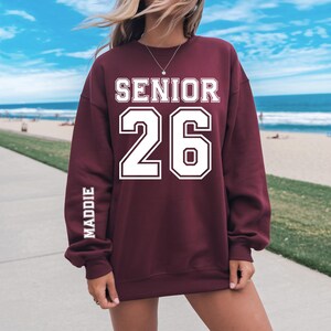 May include: Burgundy sweatshirt with the word "SENIOR" above the number "26" in white block letters. The name "MADDIE" is printed vertically on the left sleeve. The sweatshirt is a maroon color.