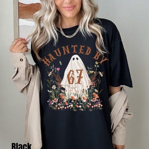 Puede incluir: Camiseta negra con el texto "HAUNTED BY" sobre un gr&aacute;fico de fantasma con el n&uacute;mero "67" y flores. La palabra "Black" est&aacute; impresa en la parte inferior de la camiseta.