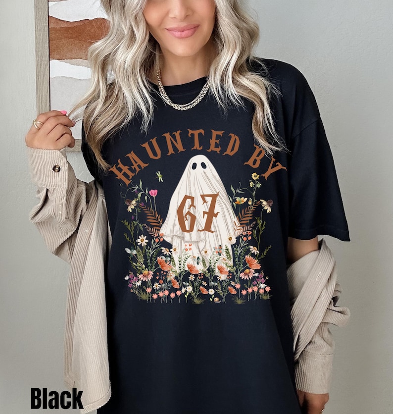 Puede incluir: Camiseta negra con el texto "HAUNTED BY" sobre un gr&aacute;fico de fantasma con el n&uacute;mero "67" y flores. La palabra "Black" est&aacute; impresa en la parte inferior de la camiseta.