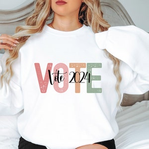 May include: White sweatshirt with the word "VOTE" in large, colorful letters with a speckled texture. The letters are pink, orange, and green. The word "Vote" is written in black script inside the "O". The year "2024" is written in black script inside the "T".