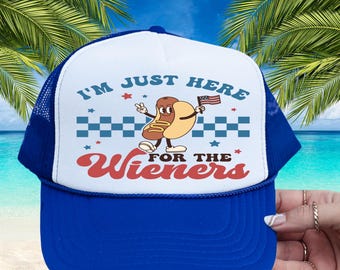 May include: A blue and white trucker hat with a graphic of a cartoon hot dog holding an American flag. The hat has the text "I'M JUST HERE FOR THE Wieners". The hat has a blue brim and mesh backing.