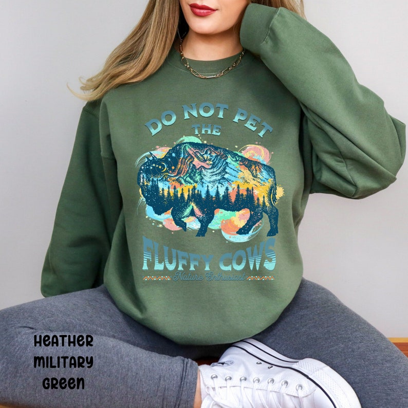 Pu&ograve; includere: Una felpa verde con una grafica di un bisonte con una scena di montagna al suo interno. Il testo sulla felpa recita "Do Not Pet The Fluffy Cows Nature's Friend".