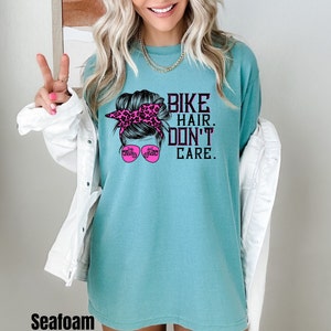 Puede incluir: Una camiseta azul turquesa con un gr&aacute;fico de una mujer con un mo&ntilde;o desordenado y gafas de sol. Las gafas de sol tienen un gr&aacute;fico de una motocicleta. El texto en la camiseta dice "Bike Hair. Don't Care."
