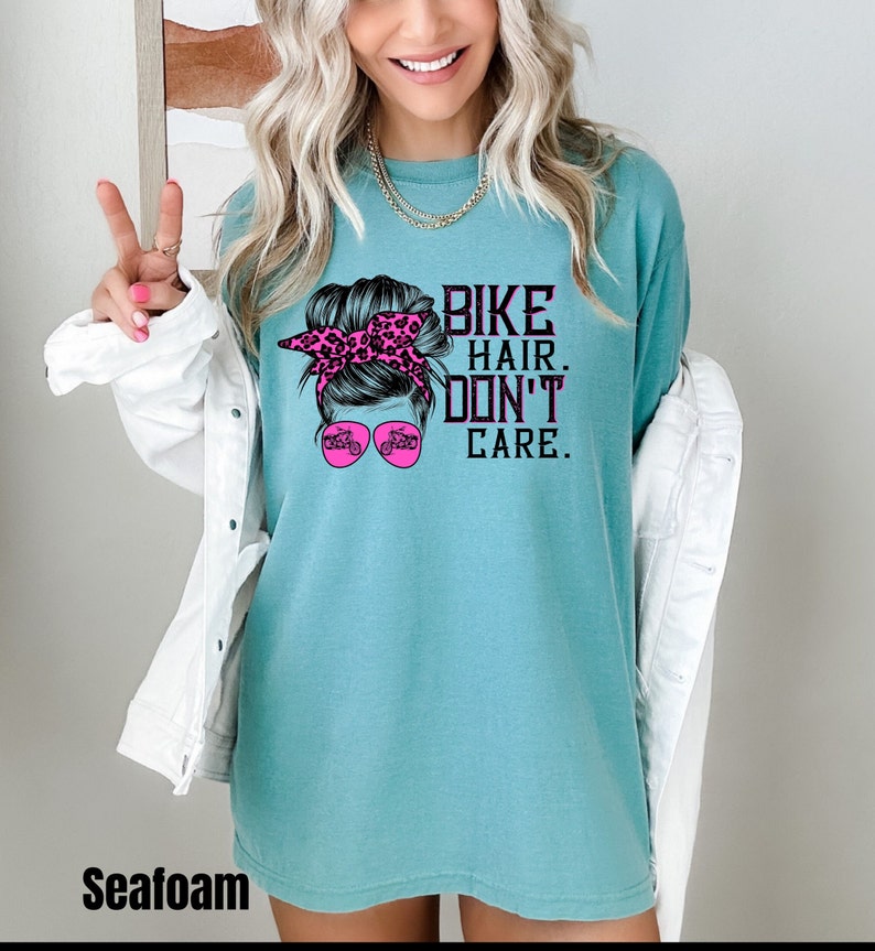 Puede incluir: Una camiseta azul turquesa con un gr&aacute;fico de una mujer con un mo&ntilde;o desordenado y gafas de sol. Las gafas de sol tienen un gr&aacute;fico de una motocicleta. El texto en la camiseta dice "Bike Hair. Don't Care."