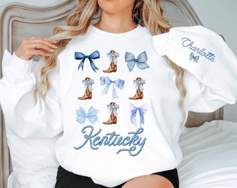 May include: White sweatshirt with a blue and brown design. The design features a repeated pattern of blue bows and brown cowboy boots. The text "Kentucky" is written in blue with a rope design. The text "Charlotte" is written in blue on the sleeve.