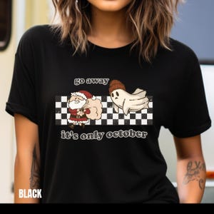 May include: Black t-shirt with a white and black checkered background. The text "go away it's only october" is printed on the shirt. A cartoon ghost wearing a hat is running away from a cartoon Santa Claus.