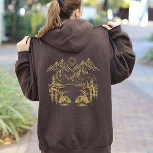 May include: A brown zip-up hoodie with a gold mountain scene on the back. The scene includes mountains, a lake, trees, and a sun.