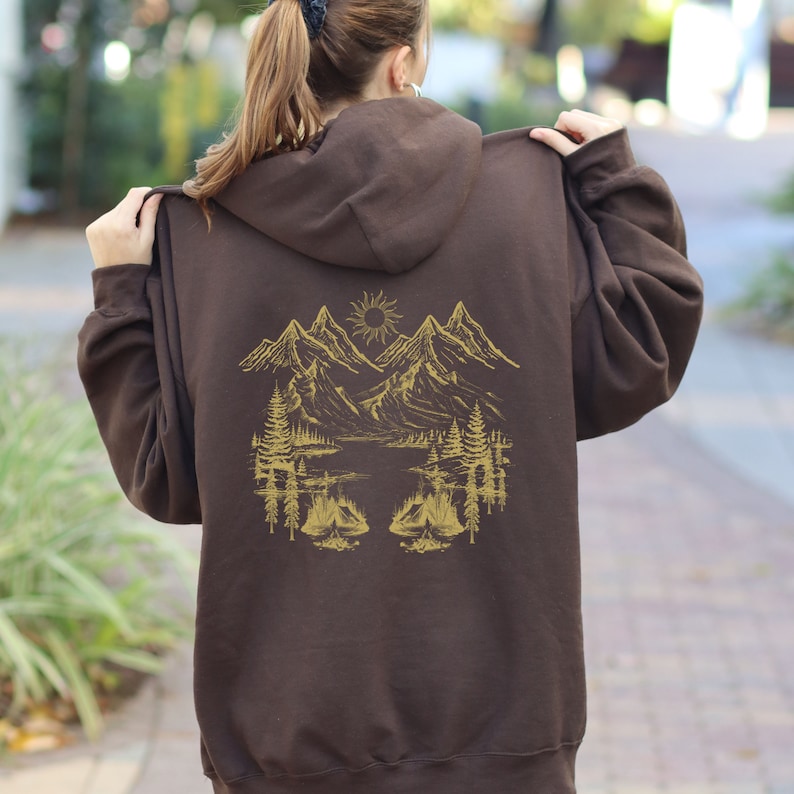 May include: A brown zip-up hoodie with a gold mountain scene on the back. The scene includes mountains, a lake, trees, and a sun.