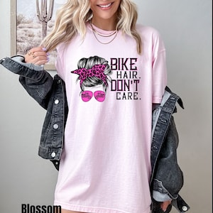 Puede incluir: Camiseta rosa con un gr&aacute;fico de una mujer con un mo&ntilde;o desordenado, gafas de sol y una motocicleta. El texto en la camiseta dice "Bike Hair. Don't Care."