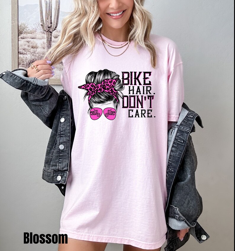 Puede incluir: Camiseta rosa con un gr&aacute;fico de una mujer con un mo&ntilde;o desordenado, gafas de sol y una motocicleta. El texto en la camiseta dice "Bike Hair. Don't Care."