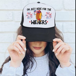 Puede incluir: Una gorra de camionero en blanco y negro con un perrito caliente rojo, blanco y azul sosteniendo una bandera. El texto en la gorra dice "I'm just here for the wieners".