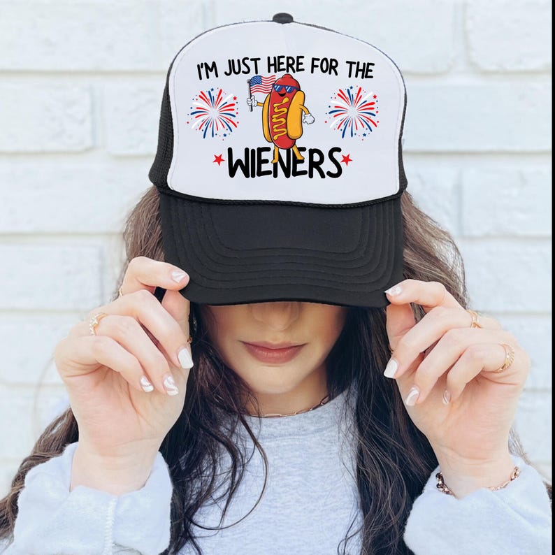 Puede incluir: Una gorra de camionero en blanco y negro con un perrito caliente rojo, blanco y azul sosteniendo una bandera. El texto en la gorra dice "I'm just here for the wieners".
