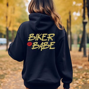 May include: A navy blue hoodie with the text "BIKER BABE" in gold glitter lettering and a red lipstick kiss graphic. The hoodie has a hood and is made of a soft material.