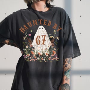 Puede incluir: Camiseta gris oscuro con el texto "HAUNTED BY 67" en letras marrones. El dise&ntilde;o presenta una figura de fantasma rodeada de flores coloridas. La camiseta es de corte holgado.