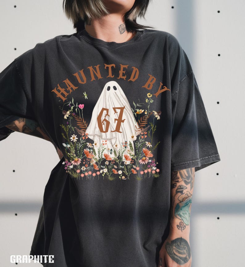 Puede incluir: Camiseta gris oscuro con el texto "HAUNTED BY 67" en letras marrones. El dise&ntilde;o presenta una figura de fantasma rodeada de flores coloridas. La camiseta es de corte holgado.