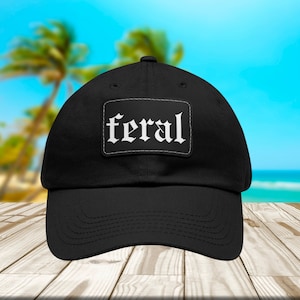 May include: Black baseball cap with the word "feral" in white gothic font on a rectangular patch. The cap is on a wooden surface with a blurred beach background.