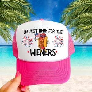 Puede incluir: Gorra de camionero rosa y blanca con un dibujo animado de un perrito caliente con gafas de sol y sosteniendo una bandera estadounidense. El texto en la gorra dice "I'm just here for the wieners".