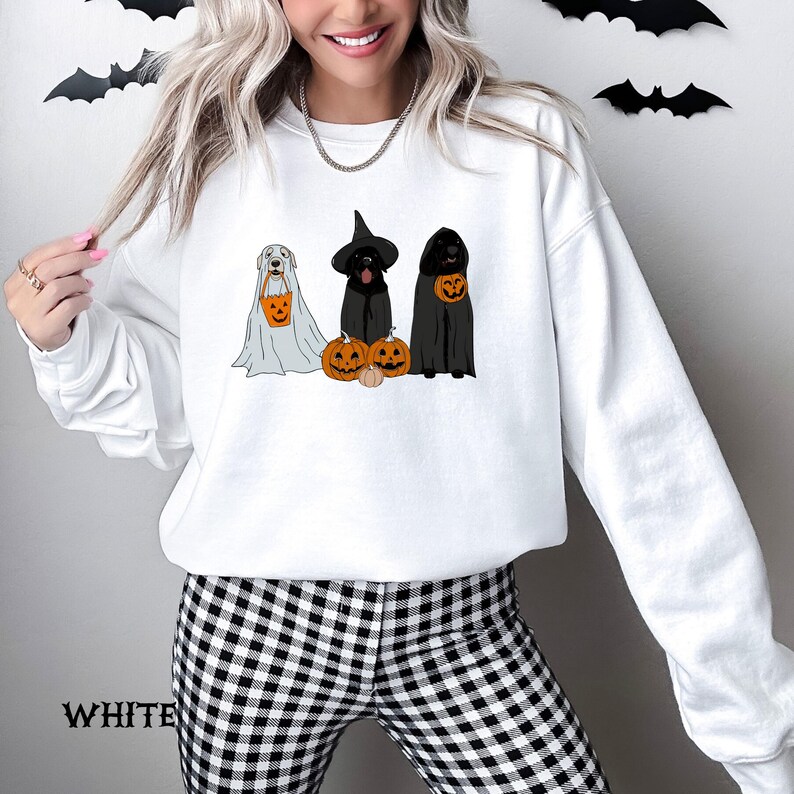 May include: White sweatshirt featuring a Halloween-themed graphic. The design includes three dogs dressed as a ghost, a witch, and a vampire, each holding a jack-o'-lantern. The word "WHITE" is printed at the bottom of the sweatshirt.