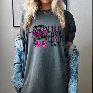 Puede incluir: Camiseta gris con un gr&aacute;fico rosa de una mujer con un mo&ntilde;o desordenado, gafas de sol y una motocicleta. El texto "Bike Hair Don't Care" est&aacute; impreso en la camiseta.