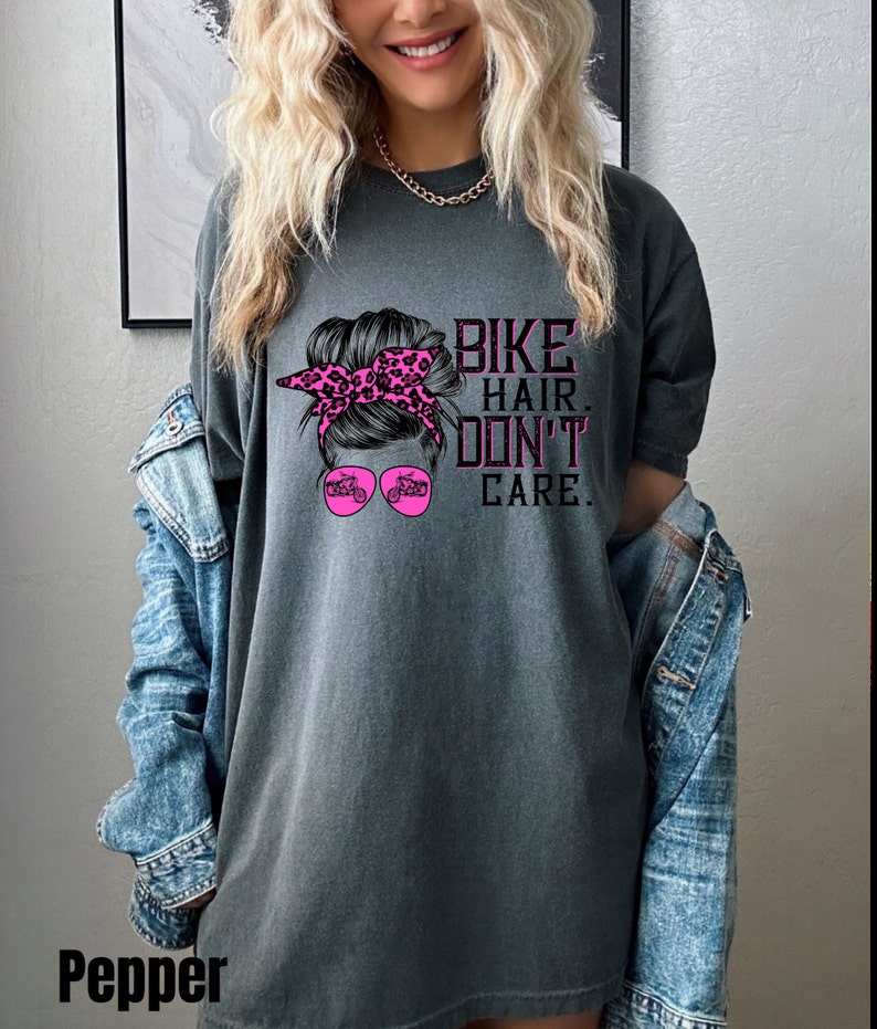 Puede incluir: Camiseta gris con un gr&aacute;fico rosa de una mujer con un mo&ntilde;o desordenado, gafas de sol y una motocicleta. El texto "Bike Hair Don't Care" est&aacute; impreso en la camiseta.