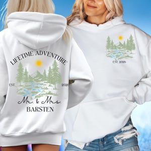 May include: White hoodie with a nature-themed design. The front features a mountain and tree graphic with the text "EST. 2026". The back displays "LIFETIME ADVENTURE" above a similar graphic, with "Mr. & Mrs. BARSTEN" below.