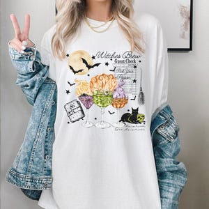 May include: White t-shirt with a graphic of witches' brew cocktails, a black cat, and a broom. The text on the shirt reads "Witches Brew Guest Check Pick Your Poison Massachusetts Salem Massachusetts".
