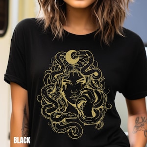 Puede incluir: Camiseta negra con un gráfico dorado de una mujer con serpientes en el pelo y una luna creciente sobre su cabeza.
