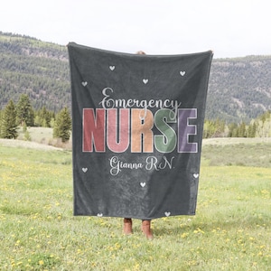 Puede incluir: Una manta de franela gris con el texto "Emergency Nurse" en colores arcoíris y el nombre "Gianna RN" en blanco. La manta se sostiene frente a una persona de pie en un campo.