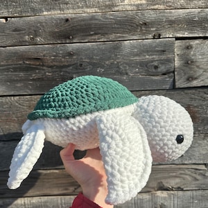 May include: A plush crochet sea turtle toy featuring a textured white body and head with a dark green shell. The turtle has two large front flippers and a small black plastic eye visible on the side of its rounded head, crafted from soft yarn.