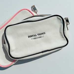 以下が含まれることがあります： 黒いジッパーの縁取りと、黒い刺繍で「DENTAL THINGS」という文字と歯科用器具の小さなアイコンが施された、長方形のオフホワイトのキャンバス地ポーチ。後ろには、ピンクのジッパーが付いた同じポーチが一部見えています。