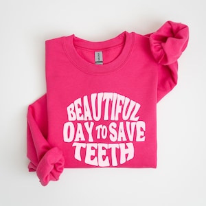 May include: A bright pink sweatshirt with the text "BEAUTIFUL DAY TO SAVE TEETH" in white, arched across the front. The sweatshirt is folded neatly, with the sleeves tucked in. The Gildan tag is visible.