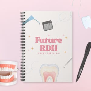 May include: A spiral-bound notebook with the text "Future RDH" in pink, along with dental-themed illustrations. A pen and a dental model are also visible. The notebook is on a pink background.