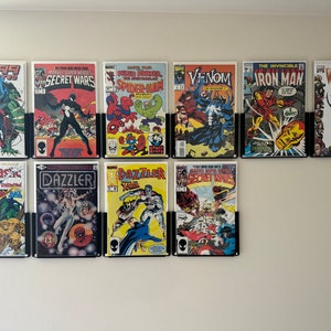 May include: A collection of ten comic books displayed on a white wall. The comic books feature colorful illustrations and titles such as "Spider-Man," "Venom," and "Iron Man." Each comic is held in a black wall mount.