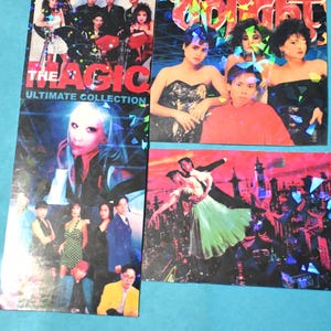 May include: A collection of vintage promotional cards with holographic accents. The cards feature images of people, text that reads "THE MAGIC ULTIMATE COLLECTION", and colorful graphics. The cards are on a blue surface.